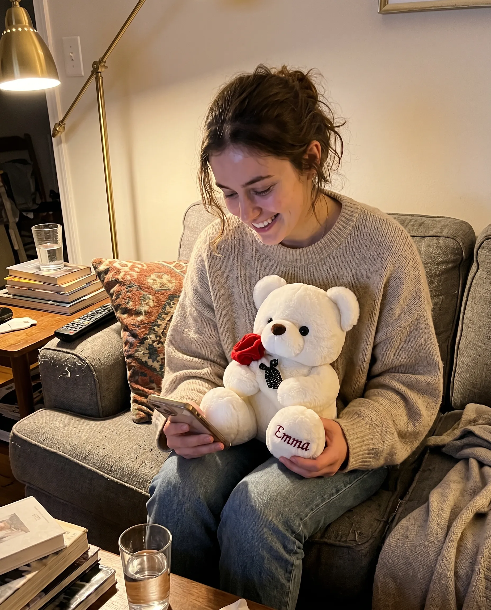 Her reaction: a young woman on the couch in warm lamp light, smiling at her phone while holding the EVR Bear with the red rose and her name embroidered on the foot